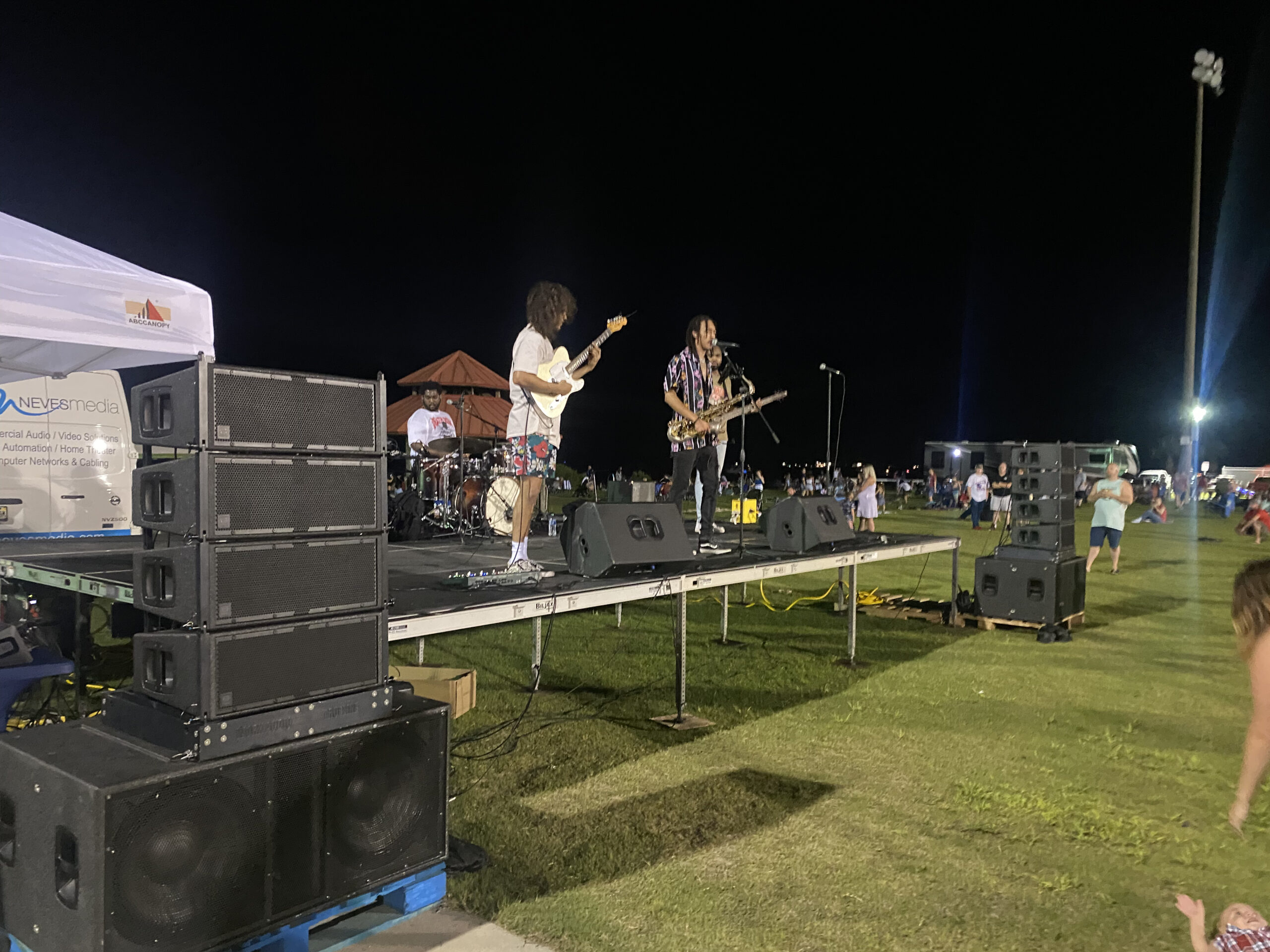 A live music performance in Panama City, FL, featuring musicians on stage, with Neves Media branding visible on a nearby tent.